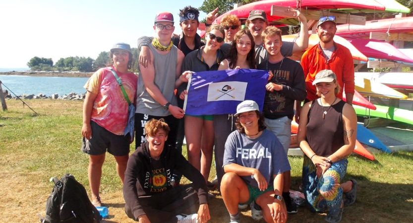 A group of people stand on grass beside a rack of kayaks and smile for a photo